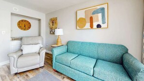 Een woonkamer met een bank, een stoel, een wandklok, een lamp en een lijst met een abstract schilderij.