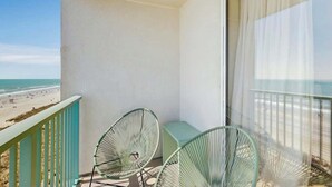 En balkong med två gröna stolar med metallställning, ett ljusblått bord och utsikt över stranden.