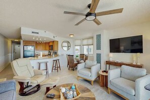 A modern living room with a ceiling fan, a television, and a dining area with a kitchen in the background. - The Pinnacle Unit 601 (North Myrtle Beach)