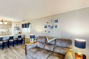 Ein Wohnzimmer mit einer Couch, einem Couchtisch und einer Lampe. Es gibt eine Küchenzeile mit einer Arbeitsplatte und Stühlen.