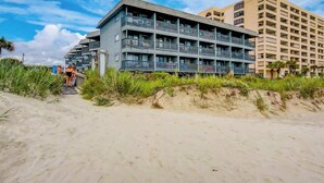 Hotel de diversos pisos amb balcons, situat a prop d'una platja sorrenca i vegetació.