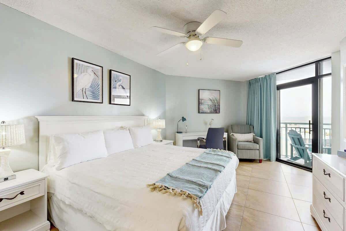 A hotel room with a bed, bedside tables, a desk, a chair, a balcony with a view, and ceiling fans.