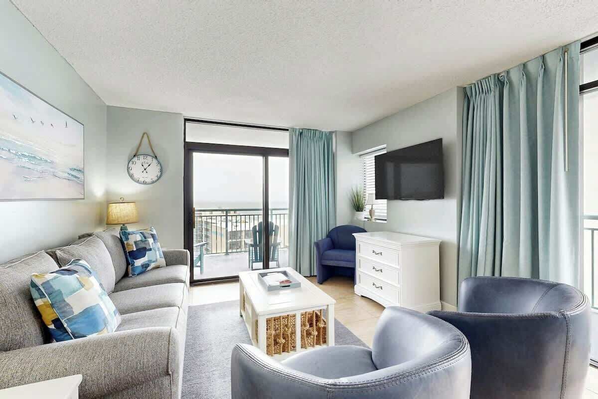 A modern living room with a sofa, armchairs, a coffee table, a flat-screen TV, and a balcony with curtains.