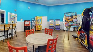 A room with a blue wall, wooden flooring, a round table with chairs, and various arcade games. - Sand Dunes Resort 910 (Myrtle Beach)