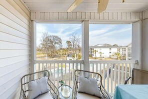 A balcony with two wicker chairs, a small table with a cup and saucer, and a view of a residential area with houses and trees. - White River Unit 31i Condo (Myrtle Beach)