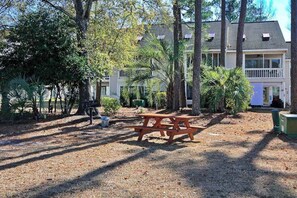 A picnic table in a shaded area with a building in the background. - Beach Golfer's Nest Retreat (Little River)