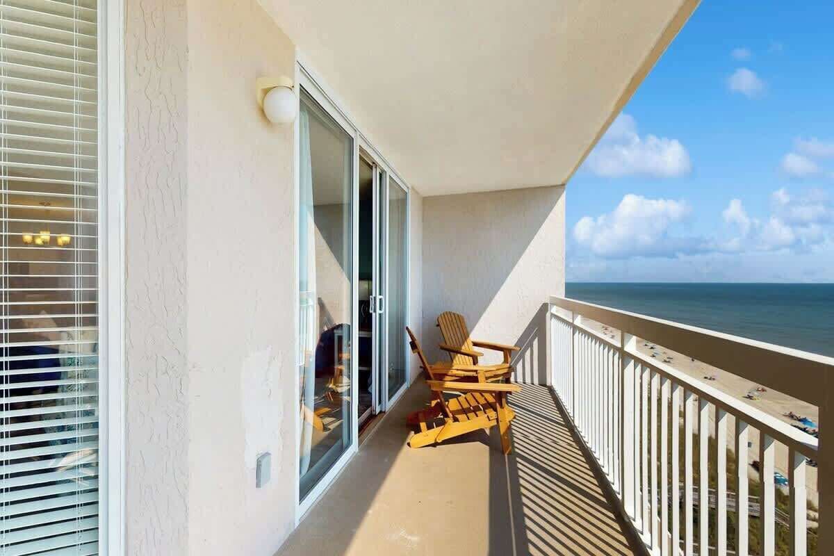 A balcony with a view of the ocean, a sliding glass door, and two wooden chairs.