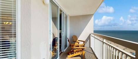 A balcony with a view of the ocean, a sliding glass door, and two wooden chairs.