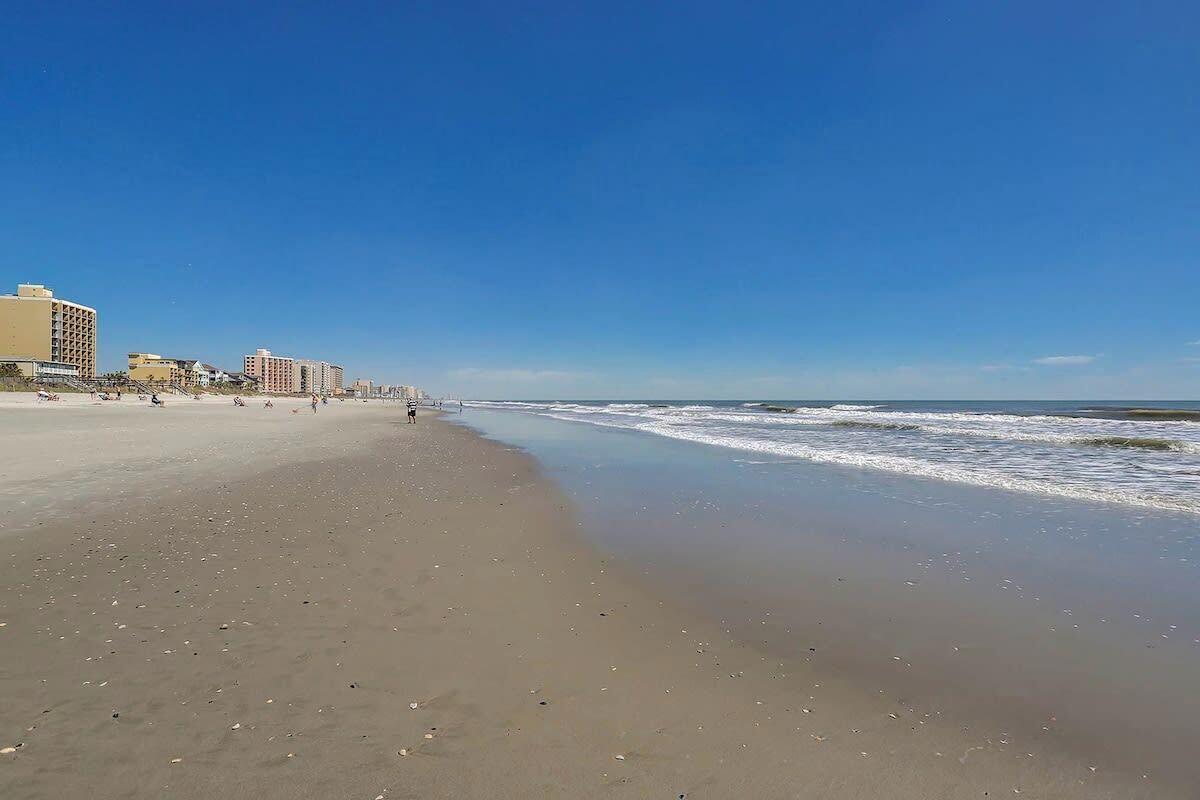 A beach with a clear sky, a sandy shore, and buildings in the background.