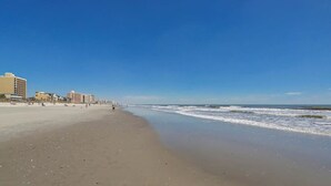 Ein Strand mit blauem Himmel, sandigem Ufer und Gebäuden im Hintergrund.
