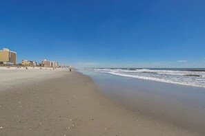 Ein Strand mit blauem Himmel, sandigem Ufer und Gebäuden im Hintergrund.
