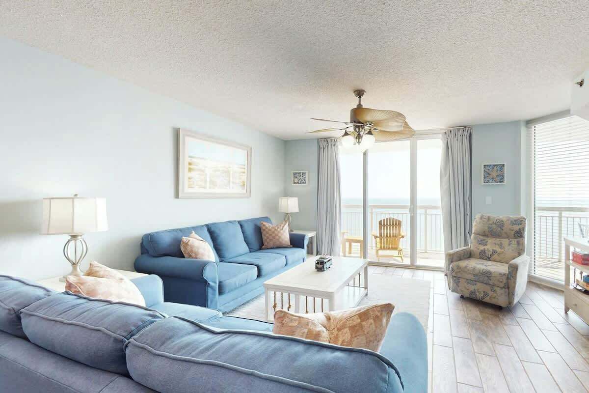 A living room with a blue sofa, a white coffee table, and a patterned armchair. There is a ceiling fan and a view of the outside through large windows.