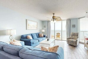 Ein Wohnzimmer mit einem blauen Sofa, einem weißen Couchtisch und einem gemusterten Sessel. Es gibt einen Deckenventilator und einen Blick ins Freie durch große Fenster.