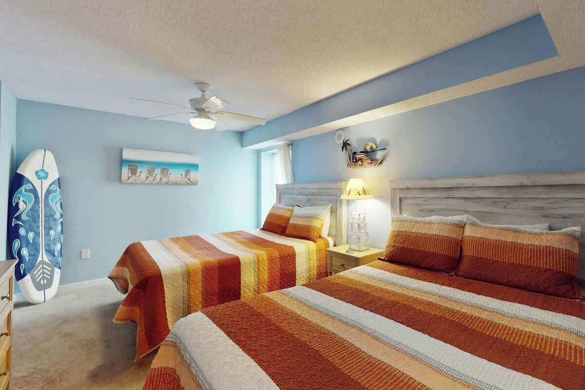 A bedroom with two beds, a ceiling fan, a surfboard, and a beach-themed wall art.