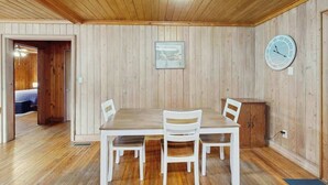 A room with a wooden ceiling, a dining table with chairs, a clock on the wall, and a door leading to another room. - The Little Brick Cottage (Murrells Inlet)