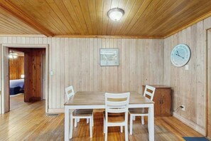 A room with a wooden ceiling, a dining table with chairs, a clock on the wall, and a door leading to another room. - The Little Brick Cottage (Murrells Inlet)