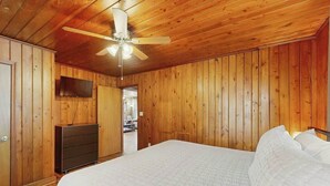 A bedroom with a bed, a ceiling fan, wooden walls, and a television. - The Little Brick Cottage (Murrells Inlet)