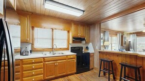 A kitchen with wooden cabinets, a black stove, and two bar stools. - The Little Brick Cottage (Murrells Inlet)