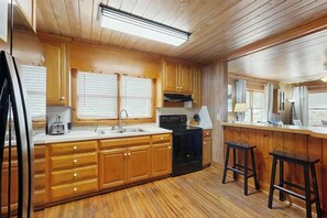 A kitchen with wooden cabinets, a black stove, and two bar stools. - The Little Brick Cottage (Murrells Inlet)