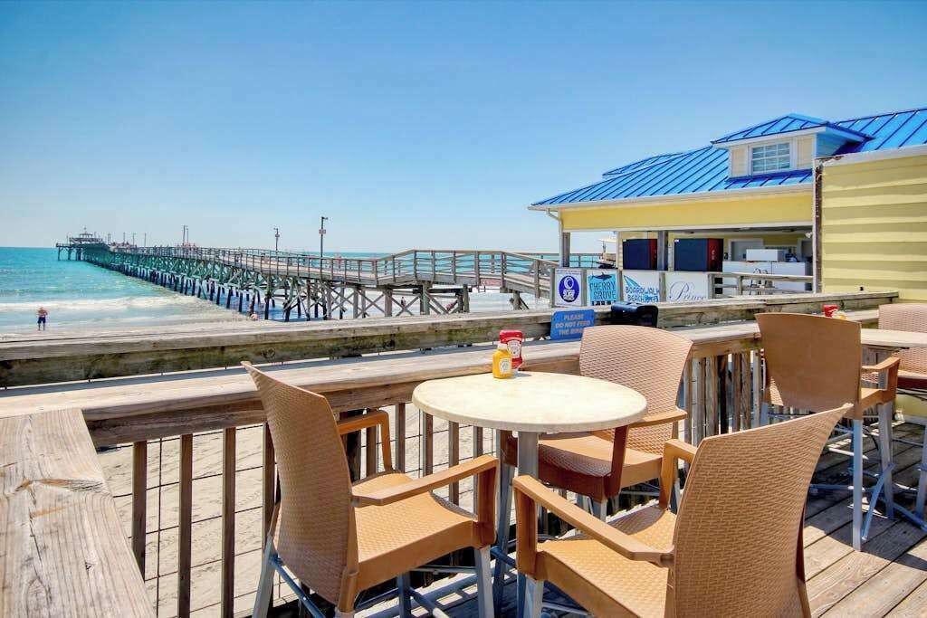 Een terras buiten met rieten stoelen en een tafel, met uitzicht op een strand en een pier.