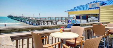 An outdoor seating area with wicker chairs and a table, overlooking a beach and a pier.