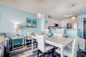 A dining area with a white table and chairs, a blue wall, and a kitchen in the background. - Prince Resort Unit 1834 (North Myrtle Beach)