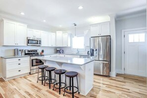 A modern kitchen with white cabinetry, a stainless steel refrigerator, a microwave, and a large island with bar stools. - Strand Ave Estate Retreat (North Myrtle Beach)