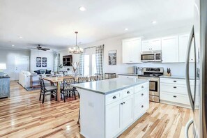 A modern kitchen with white cabinetry, a central island, and a dining area with a wooden table and chairs. - Strand Ave Estate Retreat (North Myrtle Beach)