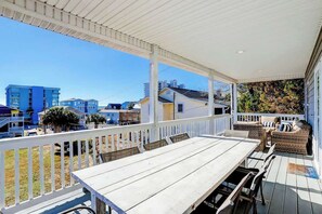 A covered outdoor deck with a dining table and chairs, a white railing, and a view of surrounding buildings. - Strand Ave Estate Retreat (North Myrtle Beach)