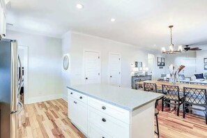 A modern kitchen with white cabinetry, a central island, and a dining area with black chairs. - Strand Ave Estate Retreat (North Myrtle Beach)