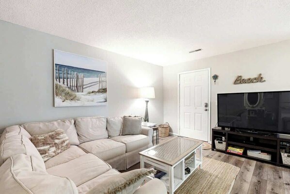 A living room with a beige sofa, a coffee table, a flat-screen TV, and a framed beach picture on the wall. - Waccamaw Condo - Unit 6 (Murrells Inlet)