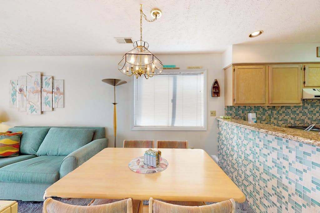 Uma sala de jantar com mesa e cadeiras de madeira, um sofá, um candelabro e uma cozinha com azulejos na parede.