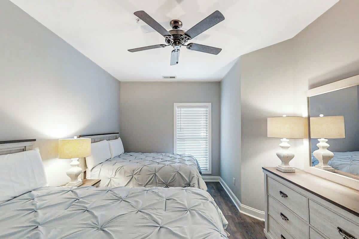 A bedroom with two beds, a ceiling fan, a dresser, and a window with blinds.