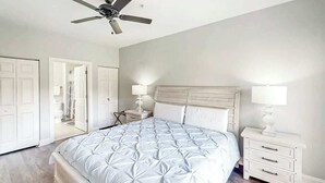 Quarto com cama, mesa de cabeceira, ventilador de teto e uma porta.