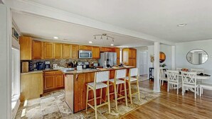 A modern kitchen with wooden cabinets, a central island with bar stools, and a dining area with a white table and chairs. - Lands End Beach Retreat (Murrells Inlet)