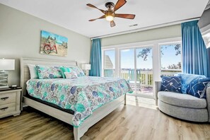 Ein Schlafzimmer mit einem Bett, einem Nachttisch, einem Deckenventilator, einem Gemälde und Blick durch eine Glastür.