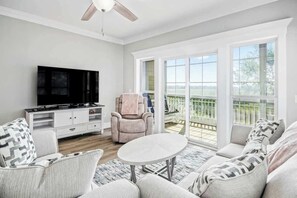 Ein Wohnzimmer mit Flachbildfernseher, einer Couch, einem Sessel und einem Tisch. Ein Balkon bietet Blick ins Freie.
