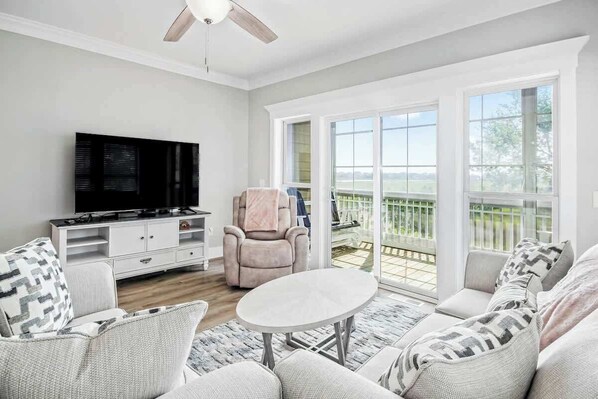 Ein Wohnzimmer mit Flachbildfernseher, einer Couch, einem Sessel und einem Tisch. Ein Balkon bietet Blick ins Freie.