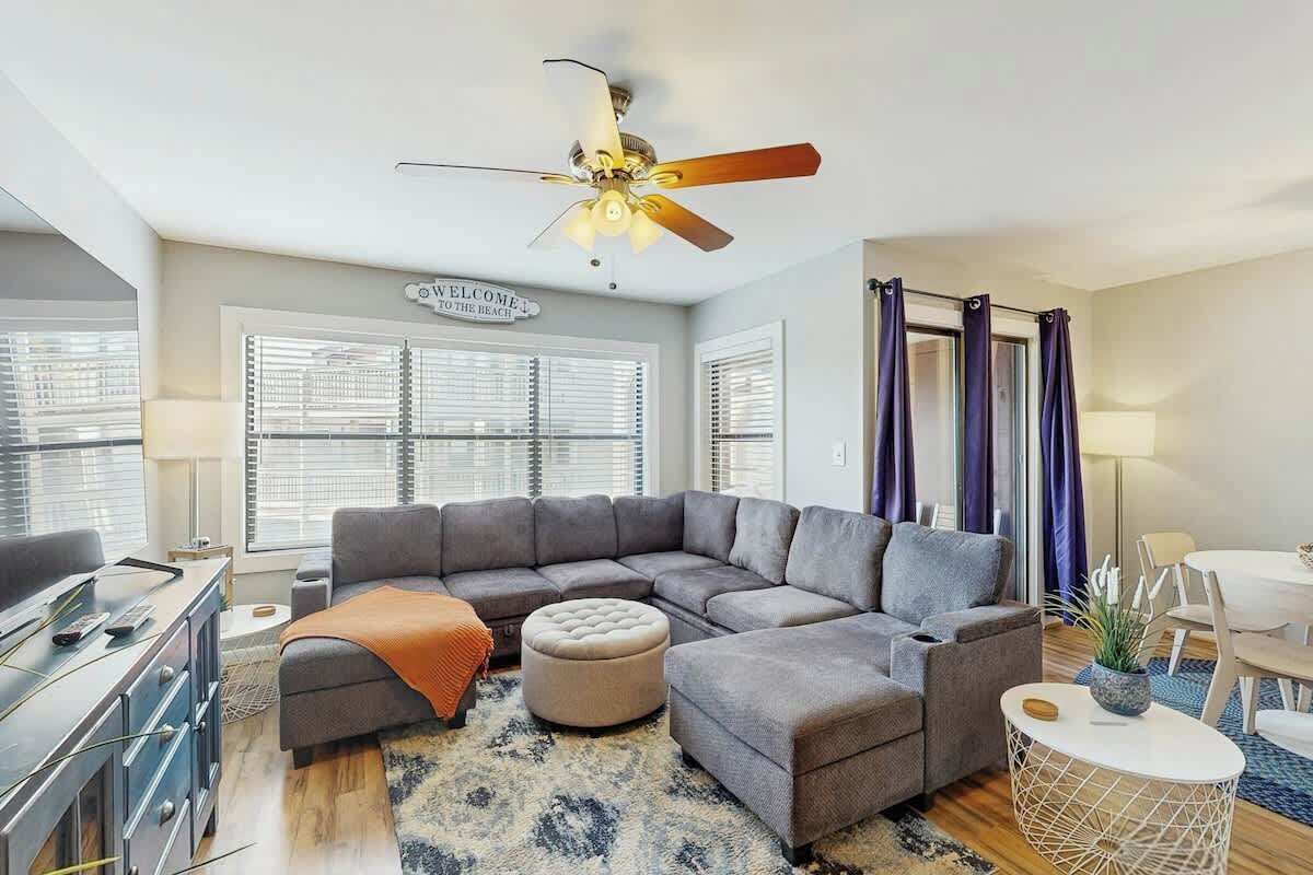 A modern living room with a sectional sofa, ottoman, ceiling fan, and a dining area with a table and chairs.