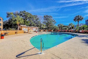 A swimming pool with a clear blue water, surrounded by a sandy area with a few trees and buildings in the background. - Dunes Pointe Unit B-11 (Myrtle Beach)