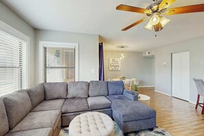 Ein Wohnzimmer mit einem Ecksofa, einem runden Hocker, einem Deckenventilator und einem Essbereich im Hintergrund.