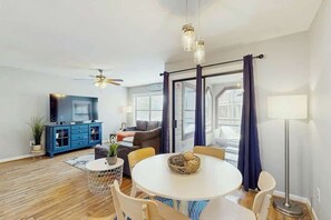 A modern living room with a round dining table, blue cabinet, flat-screen TV, ceiling fan, and large windows with curtains. - Dunes Pointe Unit B-11 (Myrtle Beach)