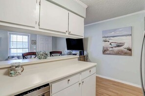 A kitchen with white cabinets, a countertop, a television, and a painting of a boat on the wall. - Toby Ct Unit A202 Condo (North Myrtle Beach)