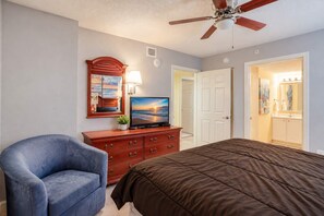 Quarto de hotel com cama, televisão, escrivaninha com gavetas, cadeira, ventoinha no teto e porta para uma casa de banho.