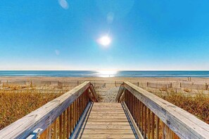 Pasarela de madera que lleva a una playa, con cielo despejado y sol.