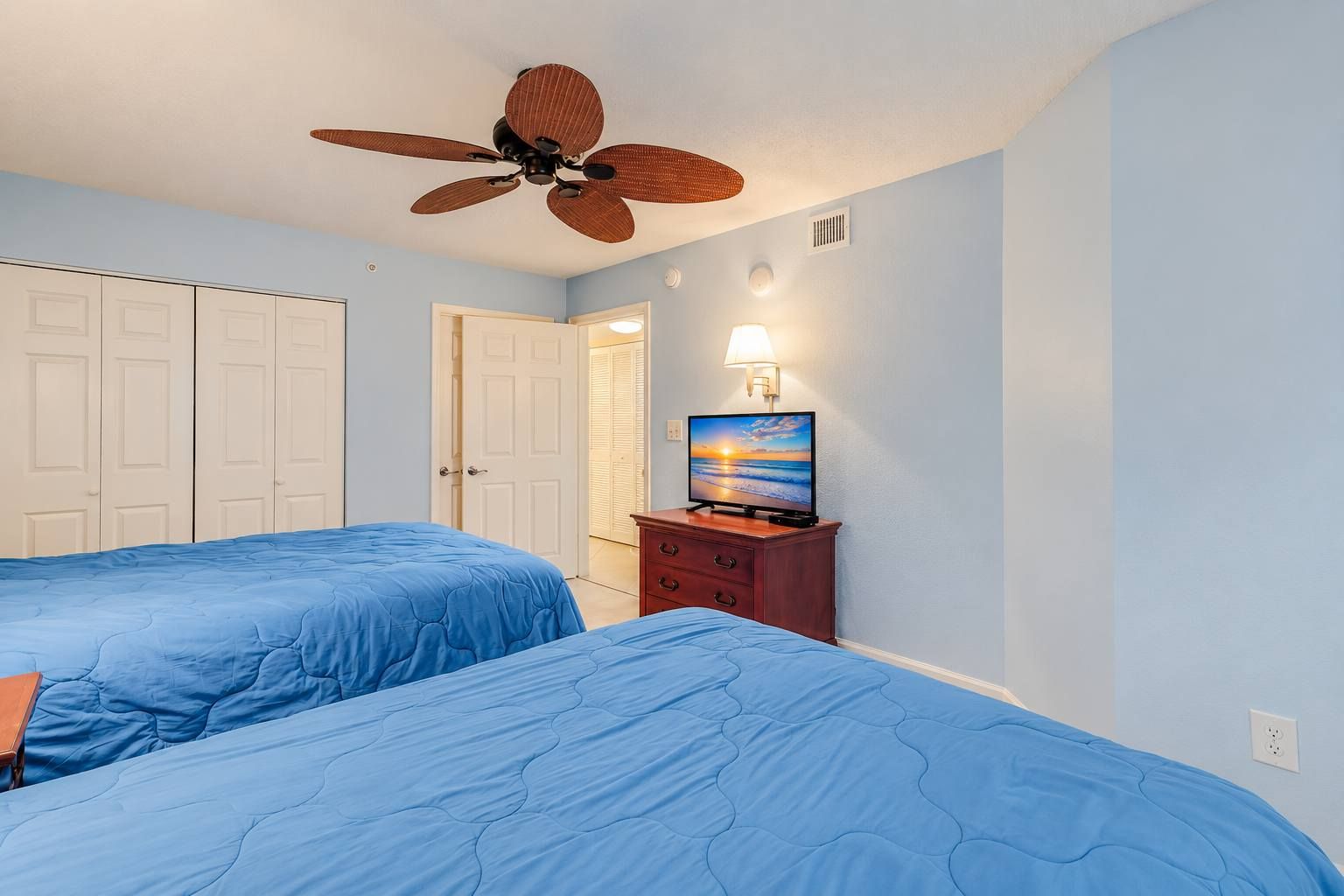 A bedroom with two beds, a ceiling fan, a nightstand with a lamp, and a TV displaying a sunset scene.