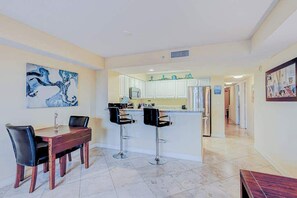 Uma área de refeições com mesa e cadeiras de madeira, uma cozinha ao fundo e um espaço bar com banquetas.
