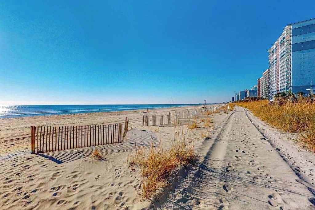 A sandy beach path with a wooden fence, leading to a beachfront with high-rise buildings in the background.