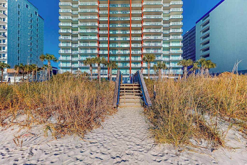 A pathway leading to a high-rise building with multiple balconies, surrounded by dry grass and palm trees.