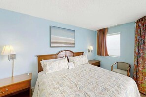 A hotel room with a bed, bedside lamps, a chair, a window with curtains, and a painting on the wall. - Roxanne Towers Unit PH1 (Myrtle Beach)